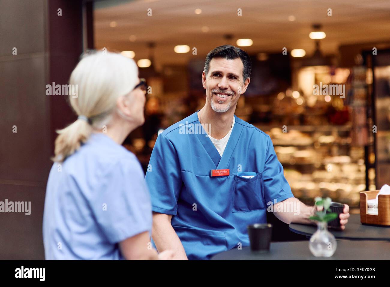 Lächelnder männlicher Gesundheitsexperte, der mit einer älteren Kollegin während der Kaffeepause in der Cafeteria des Krankenhauses spricht Stockfoto