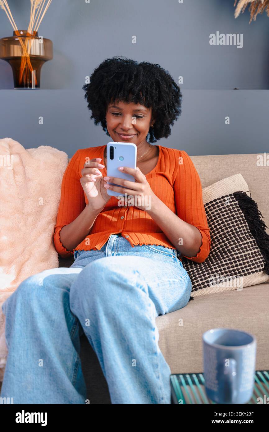 Lächelnde Frauen, die ihr Mobiltelefon auf einem bequemen Sofa in moderner Inneneinrichtung benutzen Stockfoto