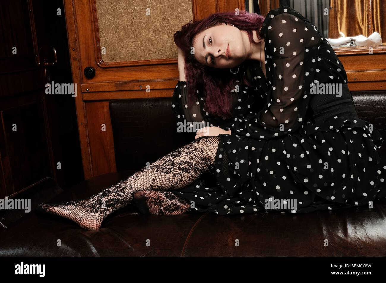 Eine anmutige Frau mit auffälligen Haaren entspannt sich auf einer klassischen Couch, verloren in Gedanken. Stockfoto