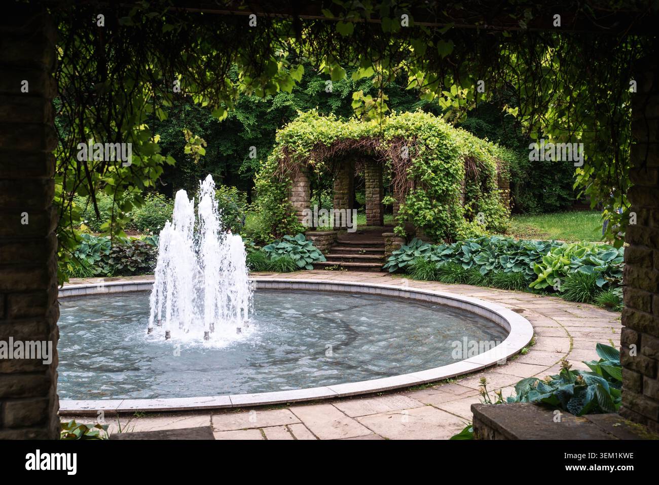 Runder Wasserbrunnen, der in einem öffentlichen Stadtpark mit einer üppig grünen Gartenpergola im Hintergrund plätschert Stockfoto
