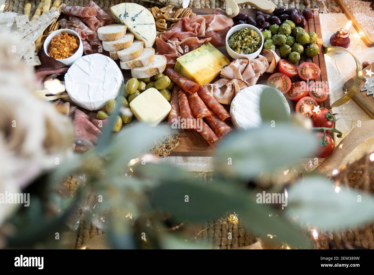 Hochwinkelblick auf ein umfangreiches Gourmet-Wurst-Weidebrett, dekoriert mit Feenlichtern, Blick durch verschwommenes Laub im Vordergrund. Stockfoto
