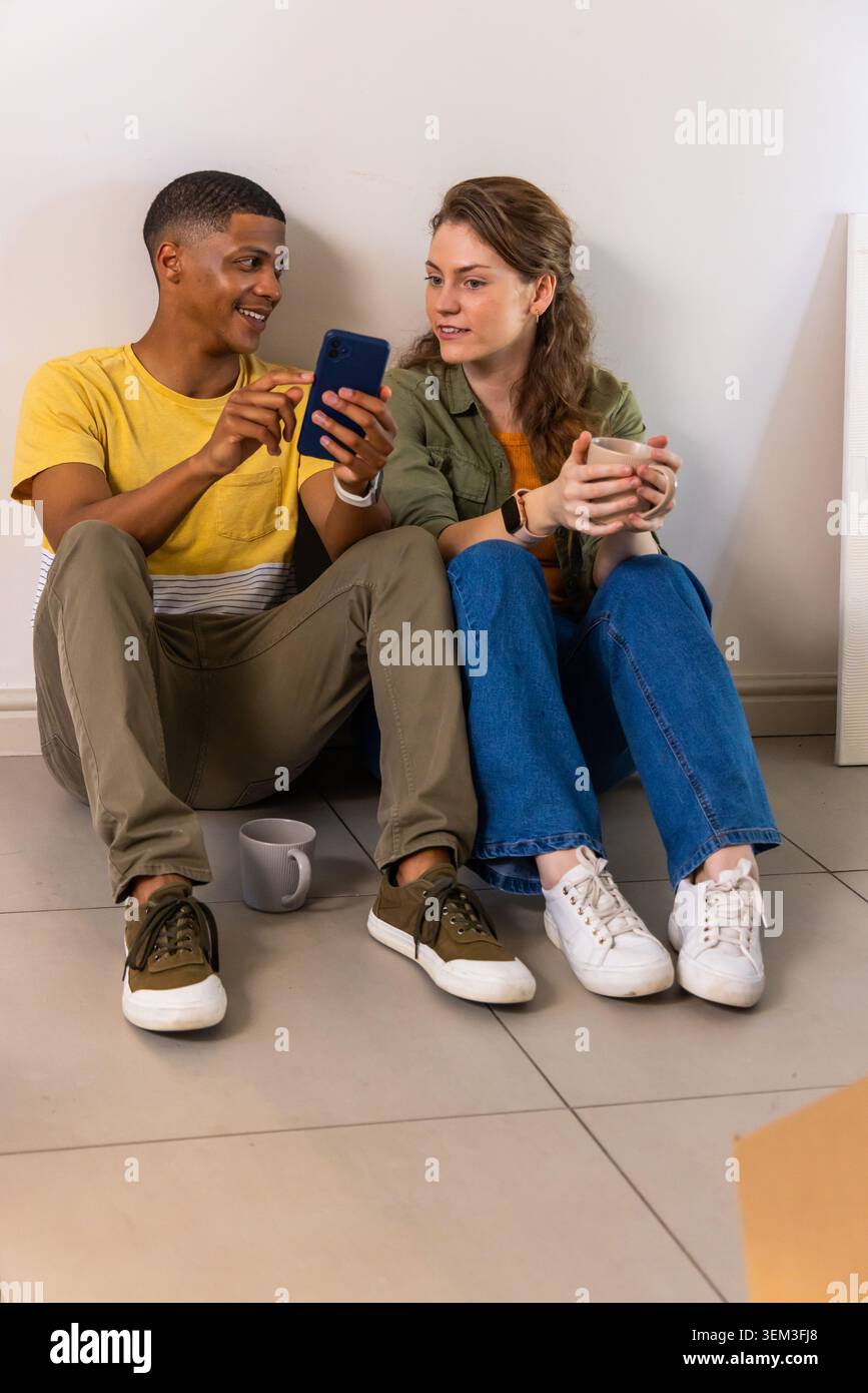 Verschiedene Paare sitzen zu Hause auf gefliestem Boden und teilen sich dunkelblaue Smartphones und Keramikbecher Stockfoto
