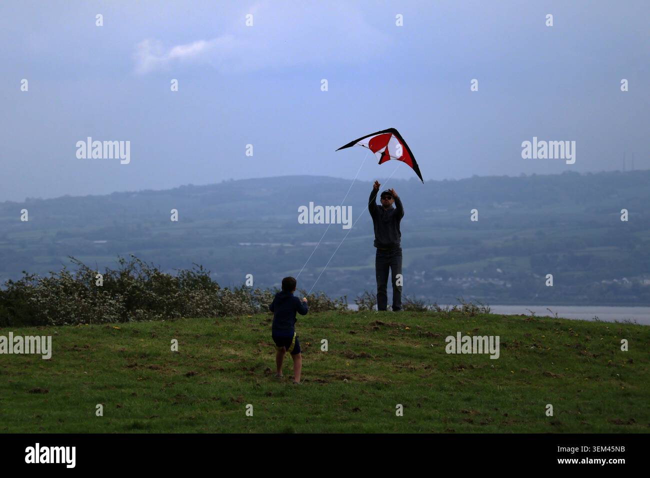 Ein Vater und sein Sohn fliegen einen rot-schwarzen Stunt-Drachen auf einem grasbewachsenen Hügel mit Blick auf die Dee-Mündung und die Küste von Nordwales in Thurstaston. Wirral, UK. Stockfoto
