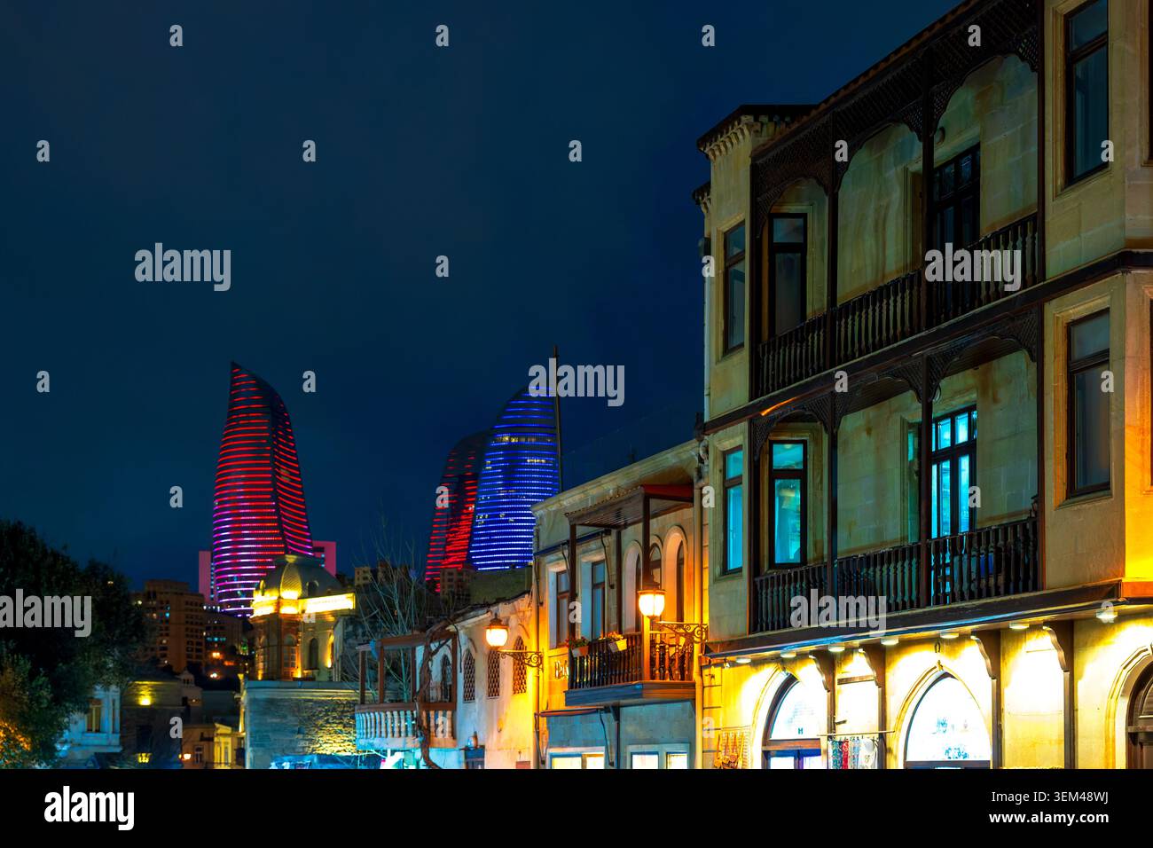 Traditionelle Steinhäuser mit Holzbalkonen in Icheri Sheher bei Nacht, mit beleuchteten Flammentürmen dahinter. Baku, Aserbaidschan. Stockfoto