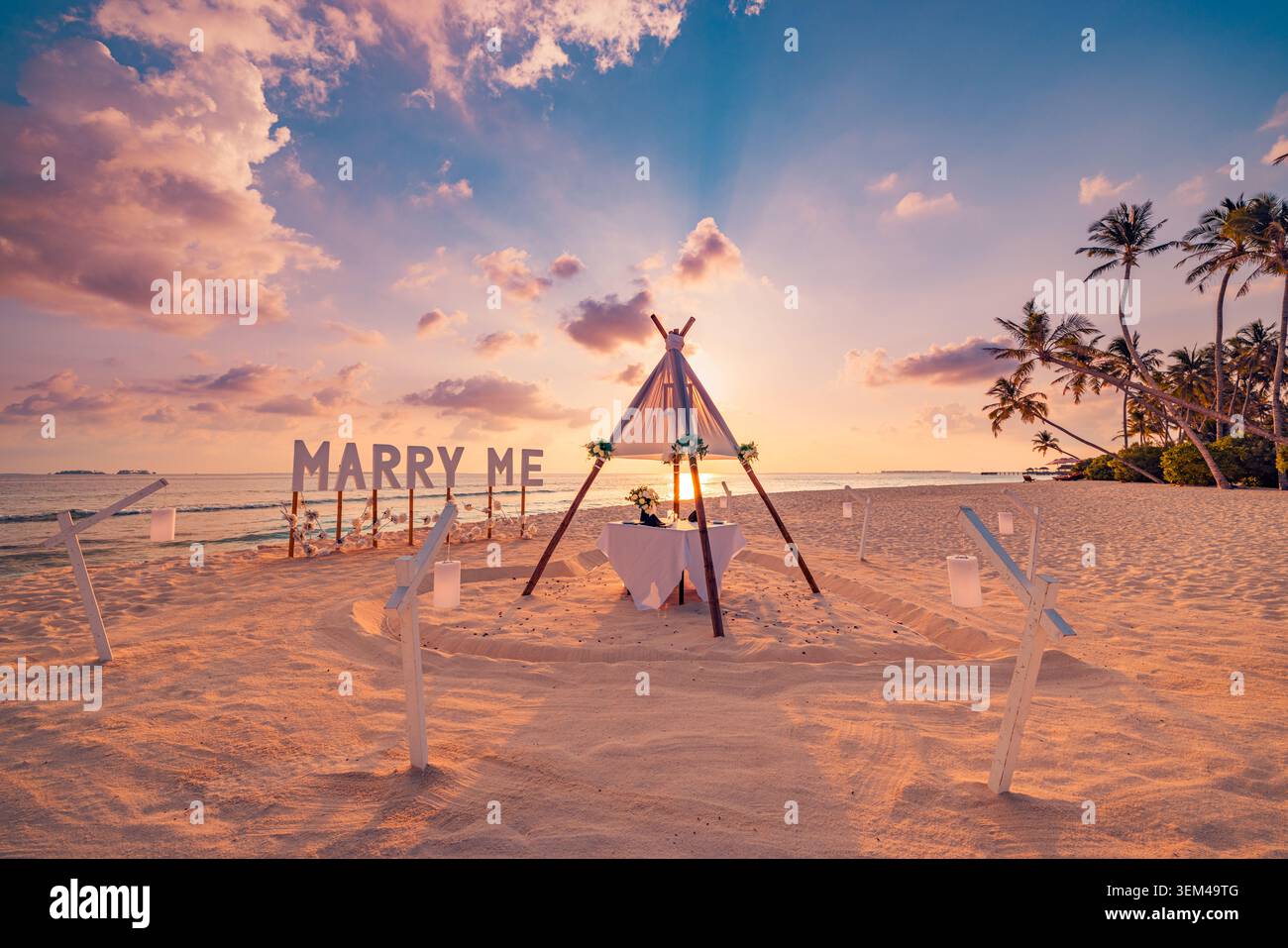 „Heirate mich“-Schild bei romantischem Strandantrag, dekorierter Esstisch unter Tipi bei Sonnenuntergang, Verlobungsfeier auf tropischem Sand, malerisch. Hochzeit Stockfoto