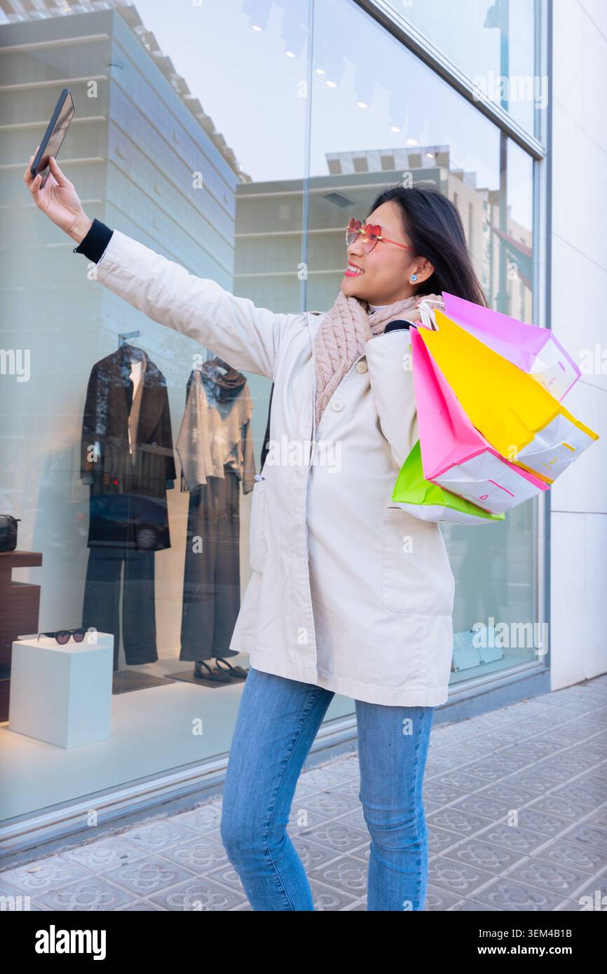 Lächelnde Frau, die Einkaufstaschen hält und draußen ein Selfie mit ihrem Smartphone macht. Stockfoto