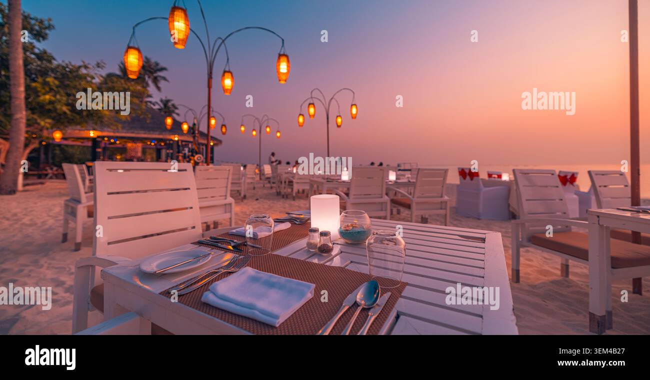 Strandrestaurant-Tische bei rotem Sonnenuntergang, leuchtende Laternen über weißen Stühlen auf dem Sand, romantisches Abendessen im Freien am Meer, tropisches Resort-Abend Stockfoto