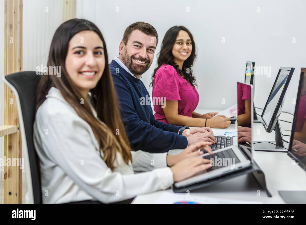 Drei Kollegen sitzen an modernen Schreibtischen in einem gut beleuchteten Coworking-Raum. Sie konzentrieren sich auf ihre Laptops, während sie an Diskussionen teilnehmen. Stockfoto
