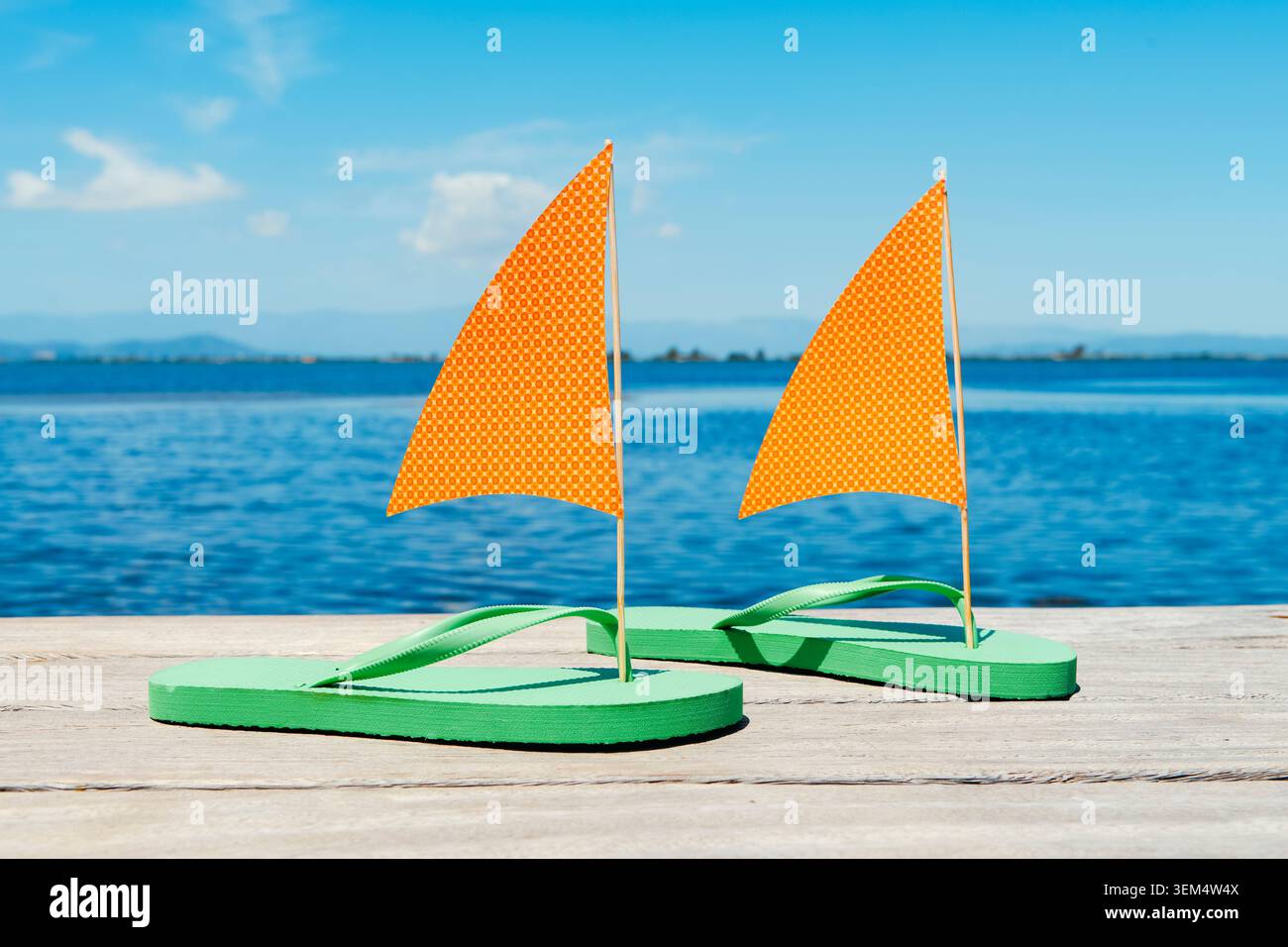 Grüne Flipflops, die in Segelboote mit orangefarbenen Papiersegeln verwandelt wurden, ruhen auf einem hölzernen Pier neben ruhigem Wasser und schaffen so ein verspieltes Sommerreisekonzept Stockfoto