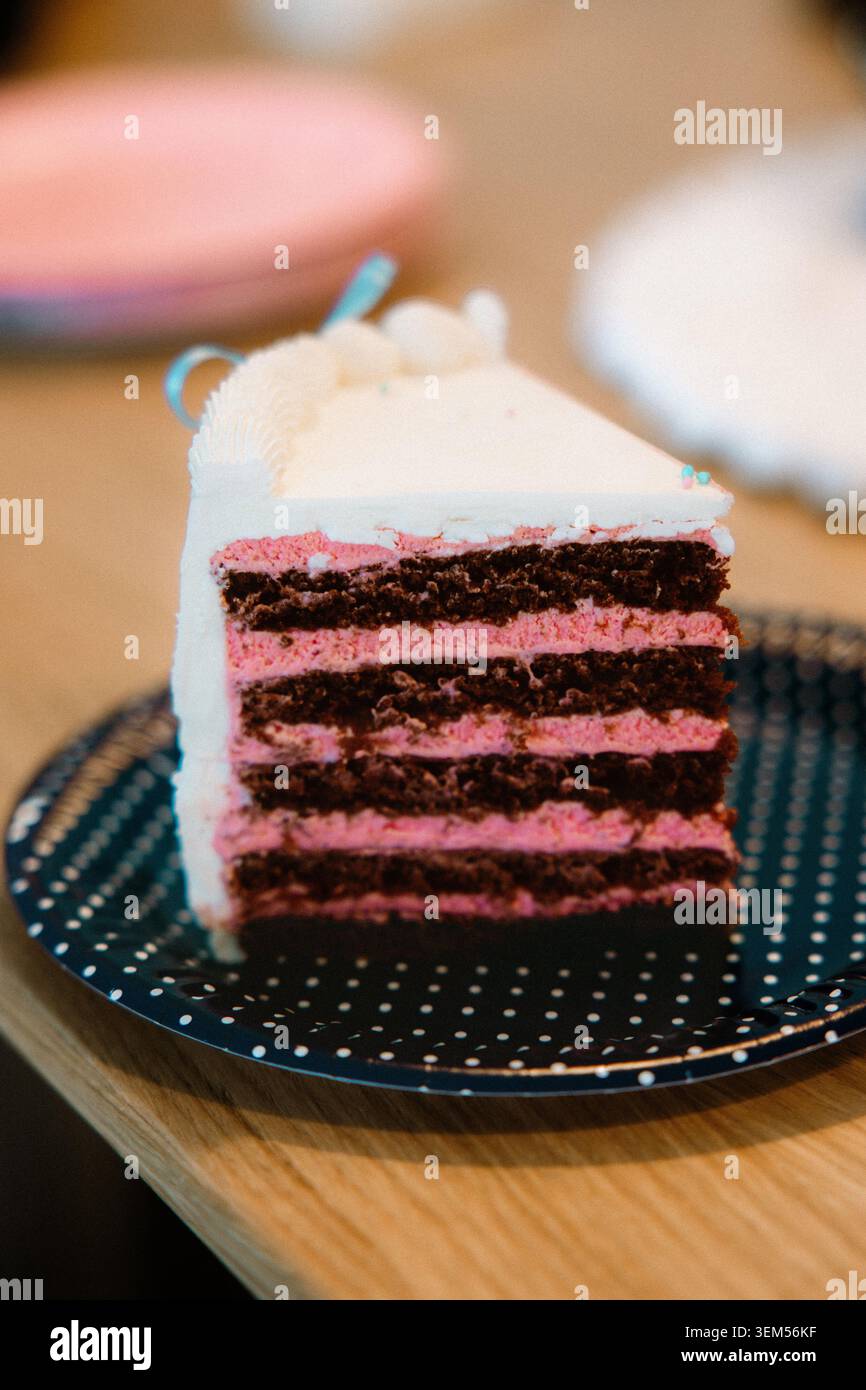 Nahaufnahme einer geschichteten Schokoladen- und Himbeercreme-Kuchenscheibe mit weißem Zuckerguss und blauem Band auf einem marineblauen Polka-Punkt Stockfoto