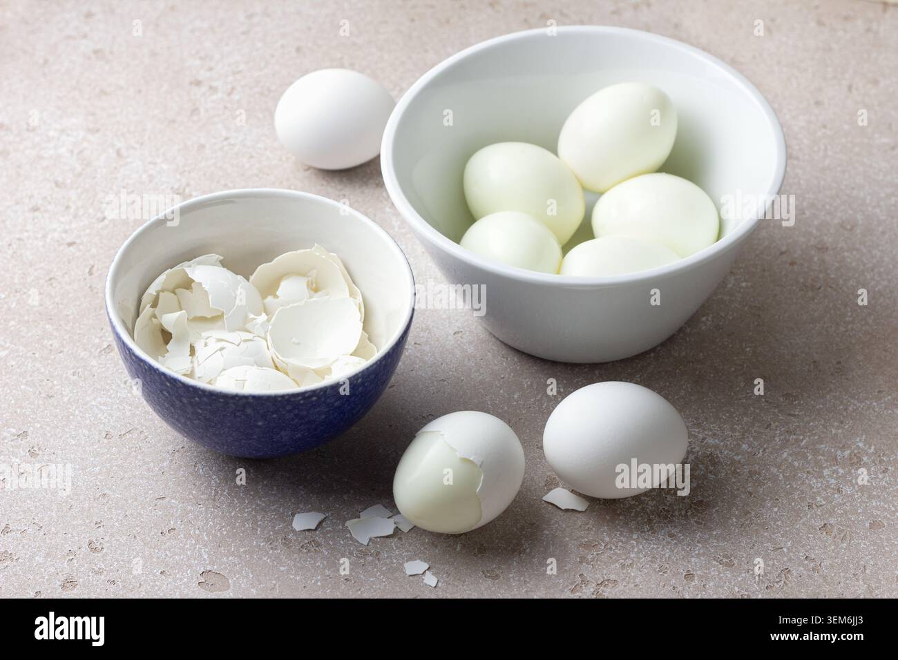 Geschälte gekochte Eier und Eierschalen in Keramikschalen auf einer hellen Steinoberfläche. Proteinreiche Lebensmittelzutat für gesunde Ernährung, Mahlzeit-Zubereitung und Stockfoto