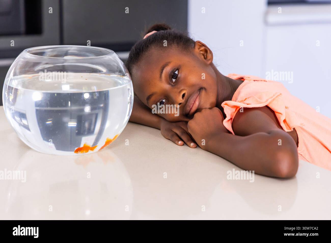 Weibliches Kind lehnt sich auf eine glänzende Arbeitsplatte in der Küche und beobachtet Fischschalen mit zwei orangefarbenen Goldfischen Stockfoto