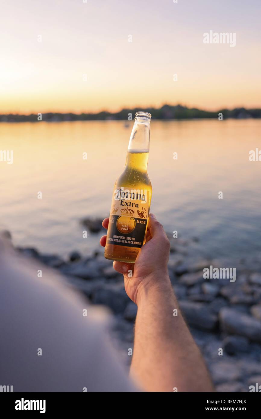 Eine Hand hält eine Bierflasche vor einem See bei Sonnenuntergang, Insel Lindau, Bodenseekreis, Deutschland Stockfoto