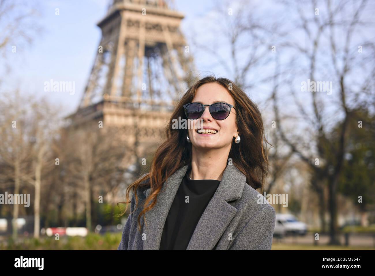 Attraktive junge Frau mit langen Haaren, Sonnenbrille und grauem Mantel, die an einem sonnigen Frühlingstag vor dem Eiffelturm lächelt Stockfoto
