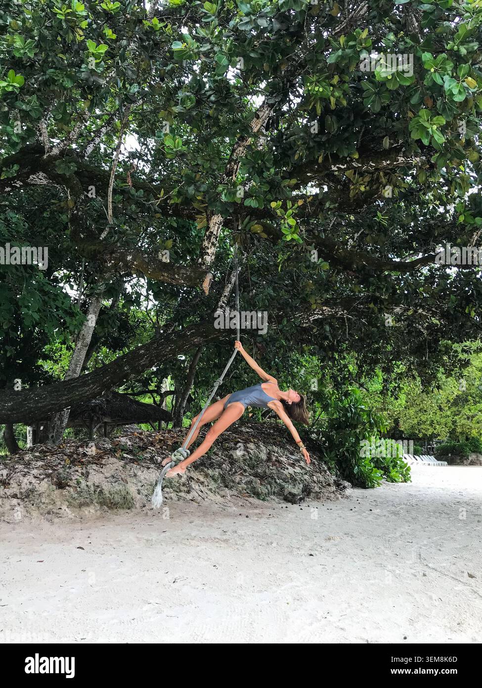 Glückliche junge Frau im Badeanzug, die an einem Seil schwingt, das an einem üppigen tropischen Baum über einem unberührten weißen Sandstrand hängt. Verspielter Urlaub. Stockfoto