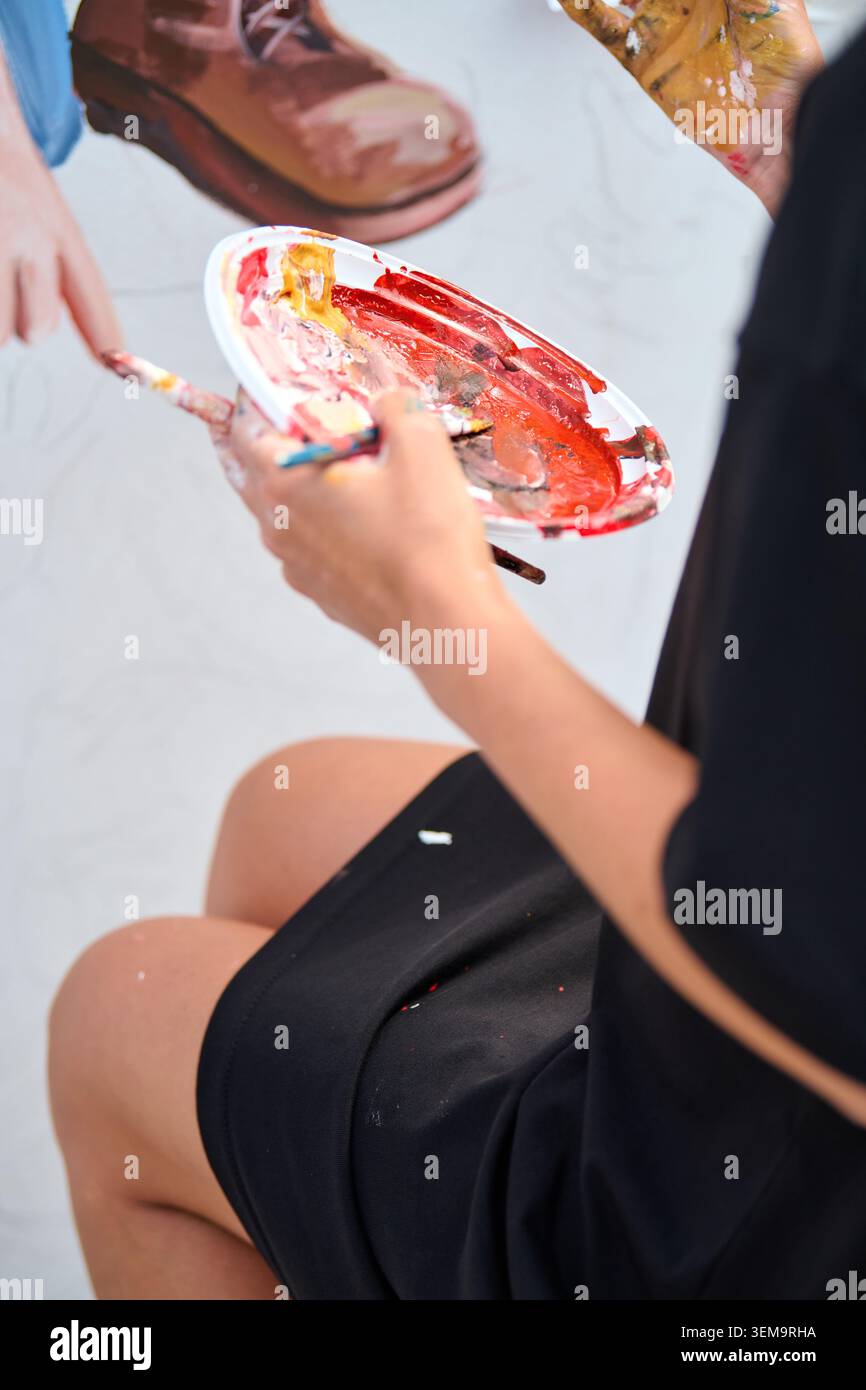 Maler, der die Farbpalette mit hellroten und gelben Acrylfarben hält und bei Tageslicht mit feinen Pinseln Striche aufträgt. Das zwanglose Ambiente konzentriert sich auf han Stockfoto