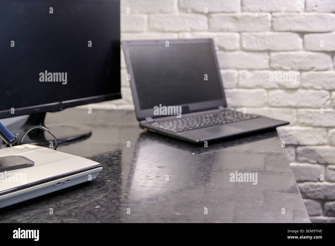 Minimalistisches Büro-Setup mit geschlossenem Laptop und ausgeschaltetem Desktop-Monitor auf reflektierender schwarzer Oberfläche mit strukturiertem weißem Backsteinhintergrund und weichem Design Stockfoto
