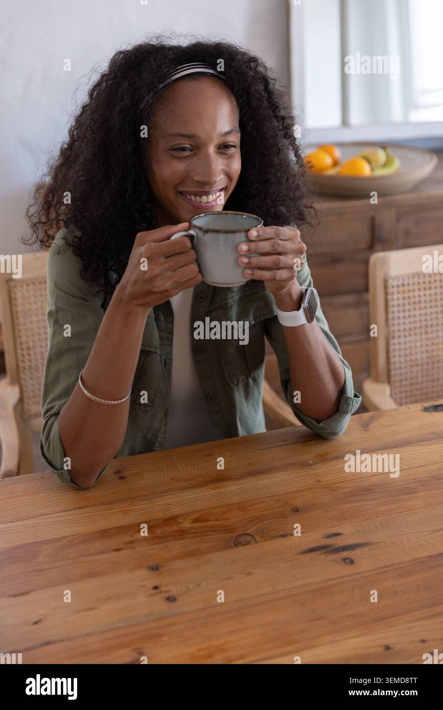 Afroamerikanerin hält einen grauen Keramikbecher und lächelt zu Hause am Esstisch in der Nähe einer Obstschale Stockfoto