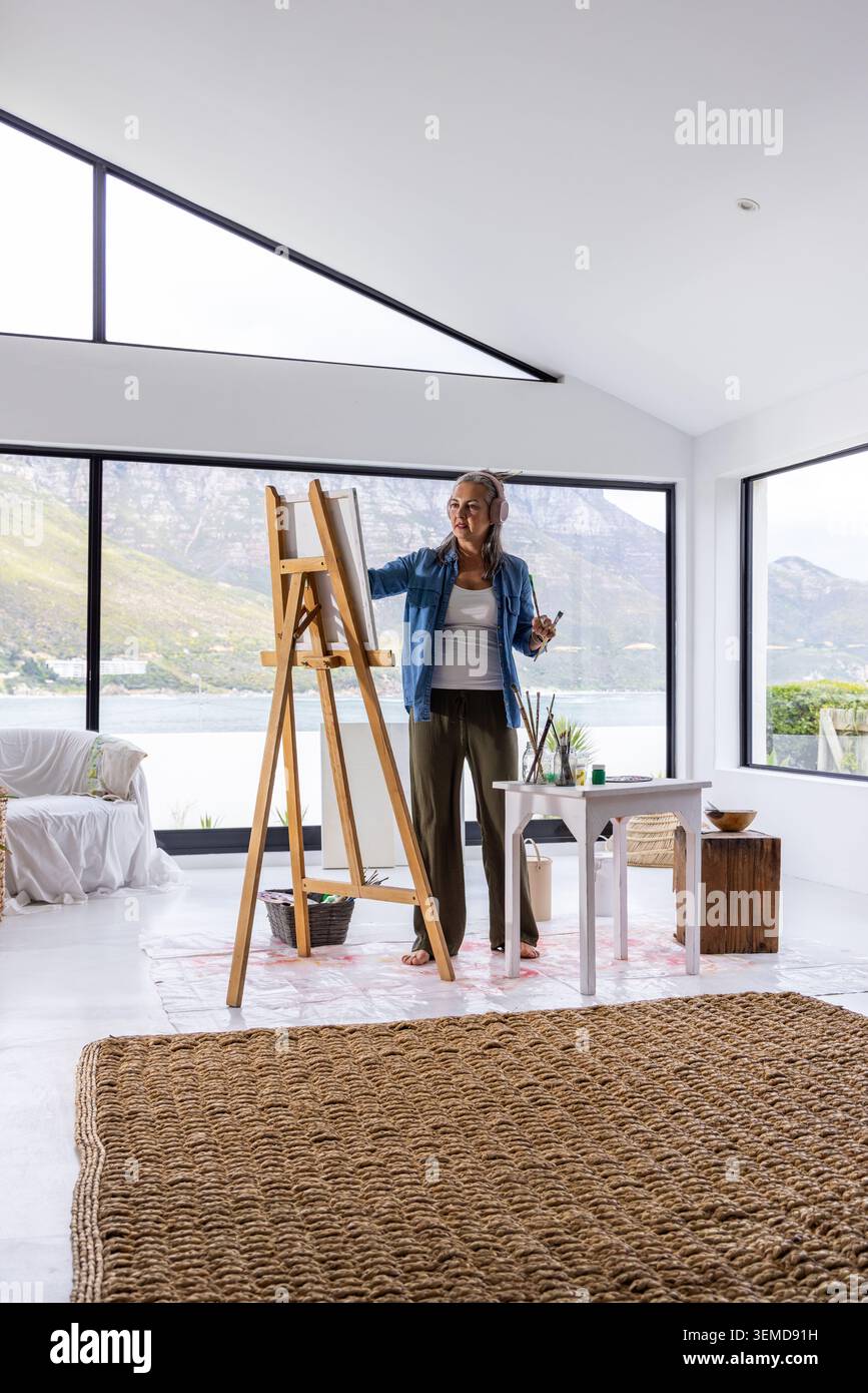 Eine Künstlerin mittleren Alters steht an einer HolzStaffelei in einem modernen Studio an der Küste und hält eine Palette, Pinsel Stockfoto