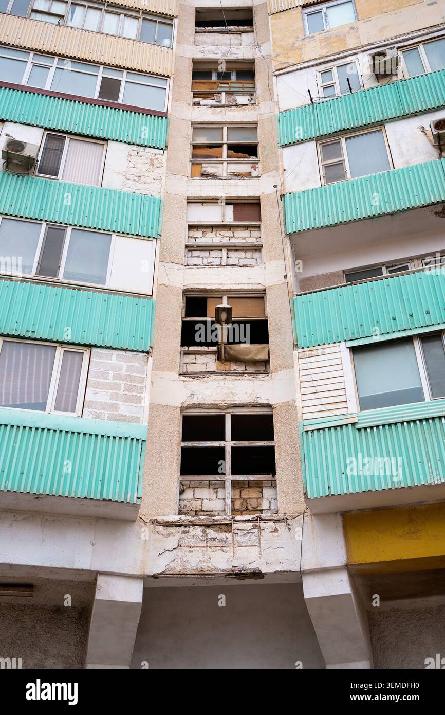 Verwitterte Wohnblockfassade aus der Sowjetzeit mit türkisfarbenen Wellbalkonen und beschädigten Fenstern, die städtische Wohnarchitektur in Uzbekis zeigen Stockfoto