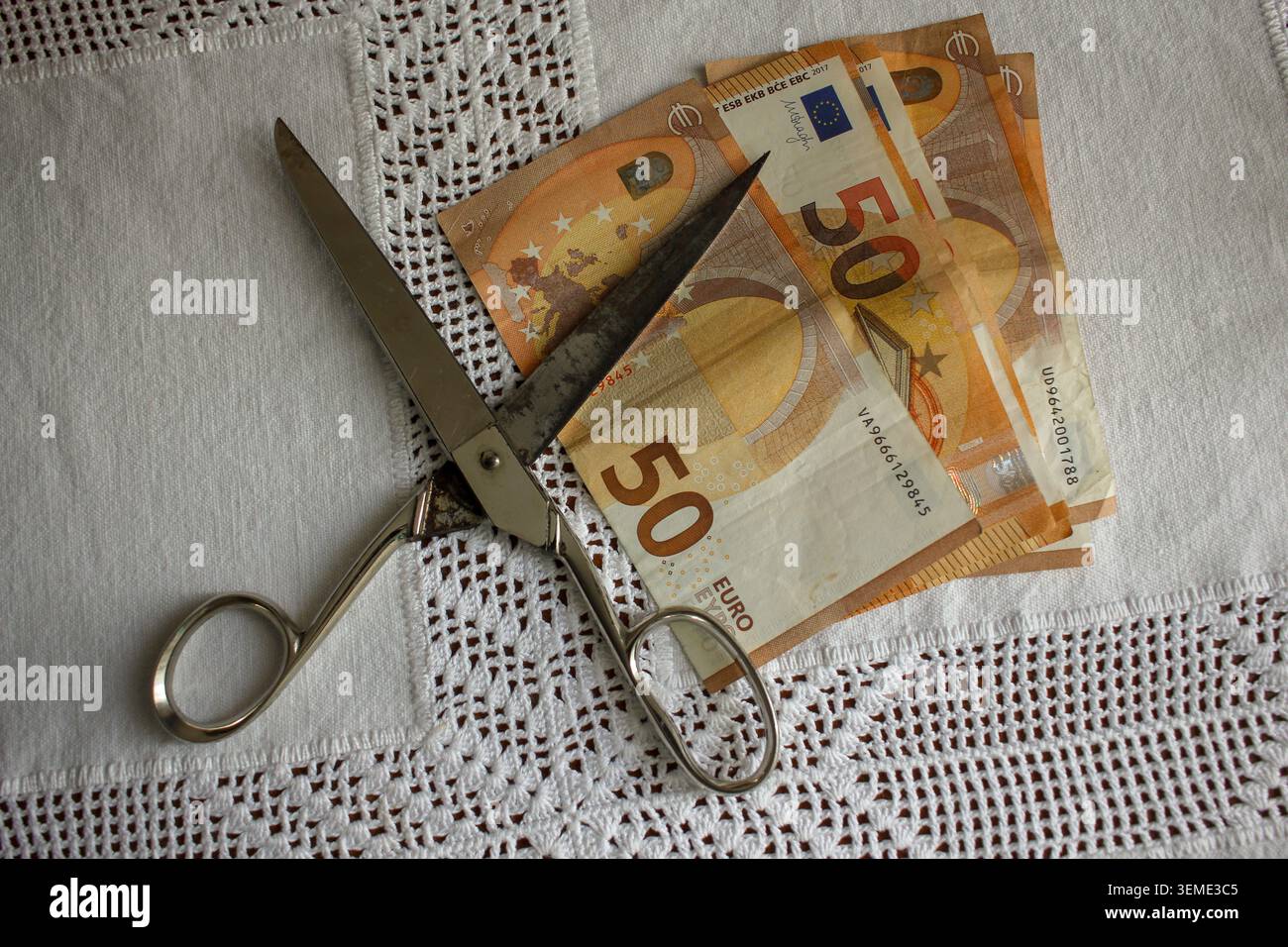 Eine Vintage-Metallschere, die über einem Fächer von 50 Euro-Scheinen auf einer weißen Tischdecke platziert wurde, symbolisiert Finanzsenkungen, Inflation und Budget o Stockfoto
