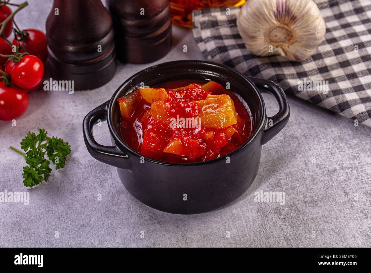 Hausgemachter Gemüseeintopf mit roten Tomaten und gelben Paprika, serviert in einer schwarzen Keramikschale mit Knoblauch, Petersilie und Holzmühlen auf grauem Steinhintergrund Stockfoto