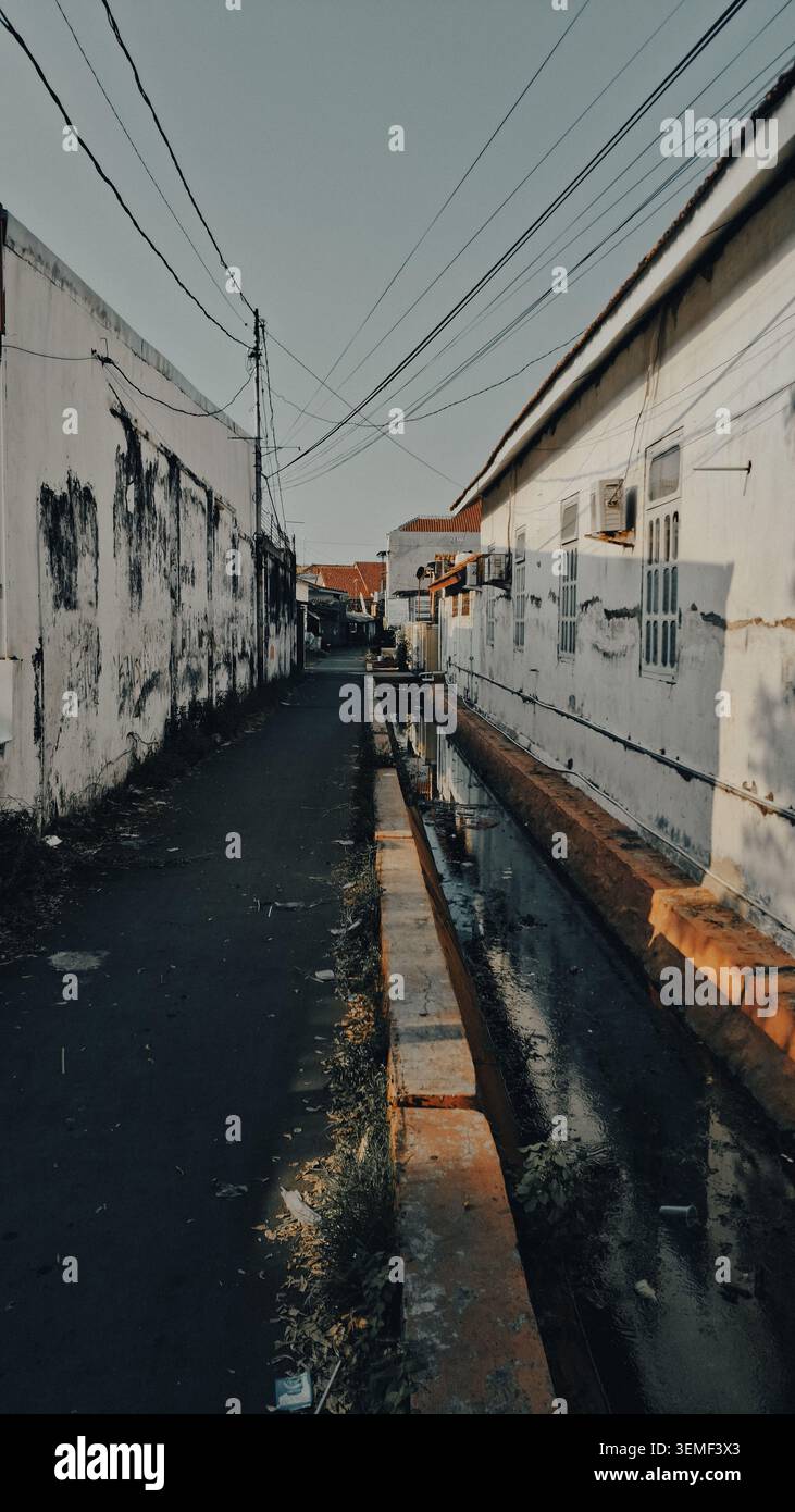 Enge Gasse in der dichten Innenstadt von Cirebon, Indonesien. Auf der einen Seite verläuft ein Kanal entlang des Weges, der von verwitterten weißen Wänden begrenzt wird Stockfoto