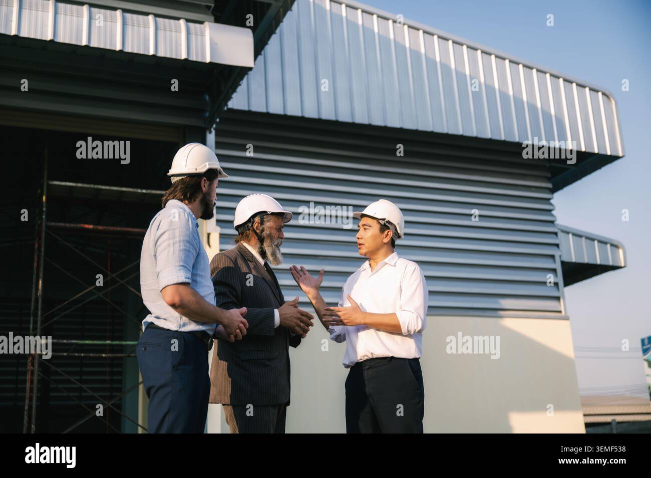 Investoreningenieure, die vor einer leeren Fabrikhalle sprechen, drei Männer tragen weiße Schutzhelme. Sie reden vor einem Metalldach Stockfoto