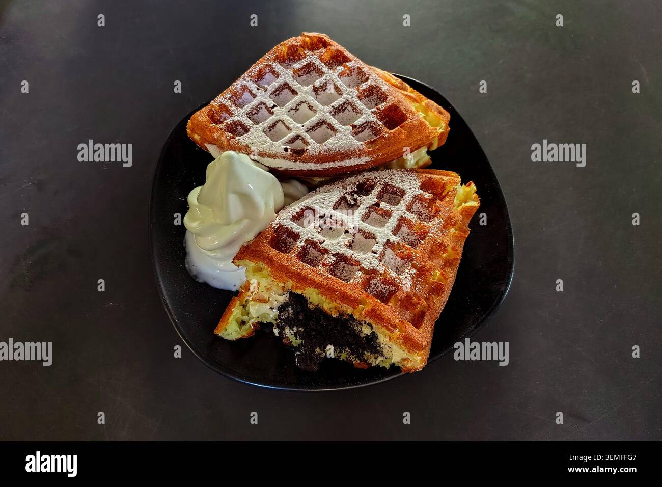 Nahaufnahme von frisch gebackenen goldenen Waffeln, gefüllt mit zerquetschten Schokoladenkeksen und Sahne, mit Puderzucker bestäubt, serviert mit weichem Vanilleeis auf einem schwarzen Teller auf dunklem Hintergrund. Stockfoto