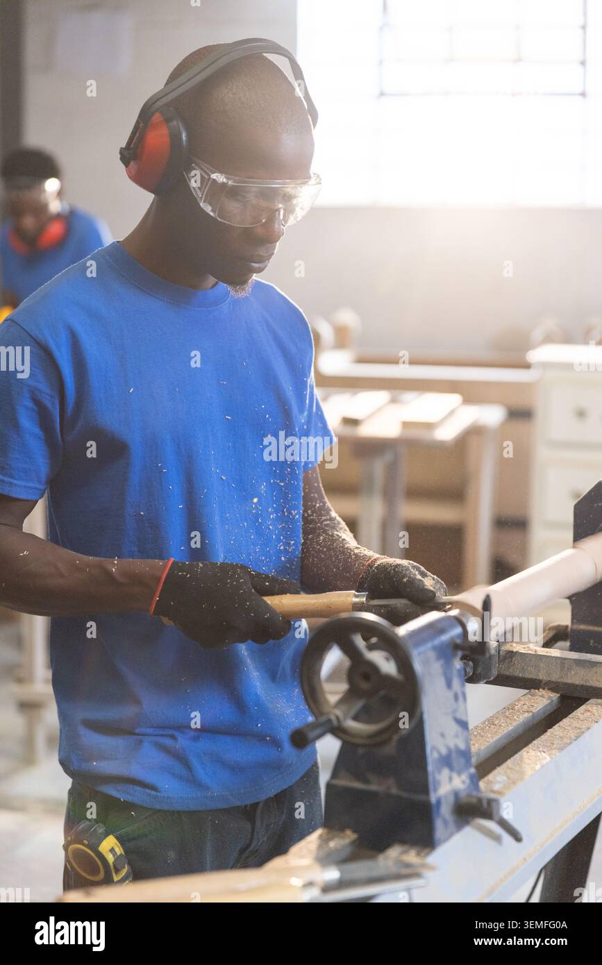 Asiatischer Mann, der Holz auf Drehwerkzeug in der Werkstatt zwischen Holzspänen mit Schutzbrille und Ohrenschützern schnitzt Stockfoto
