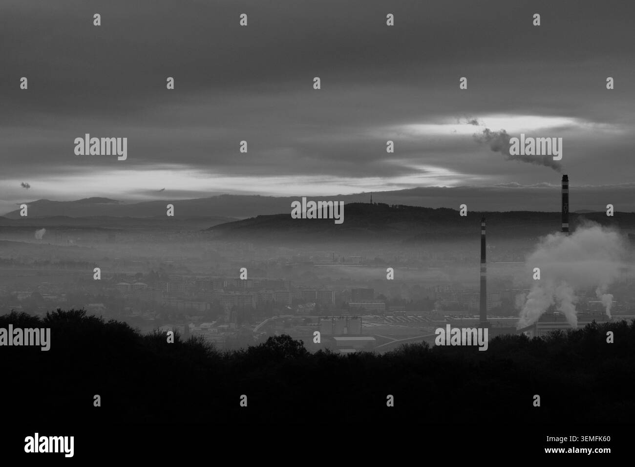 Industrieller Kamin mit Rauch, der über einer nebeligen Stadt in Schwarz-weiß aufsteigt, vom Hügel Zlutavsky kopec über Otrokovice, Tschechische Republik aus gesehen. Stockfoto