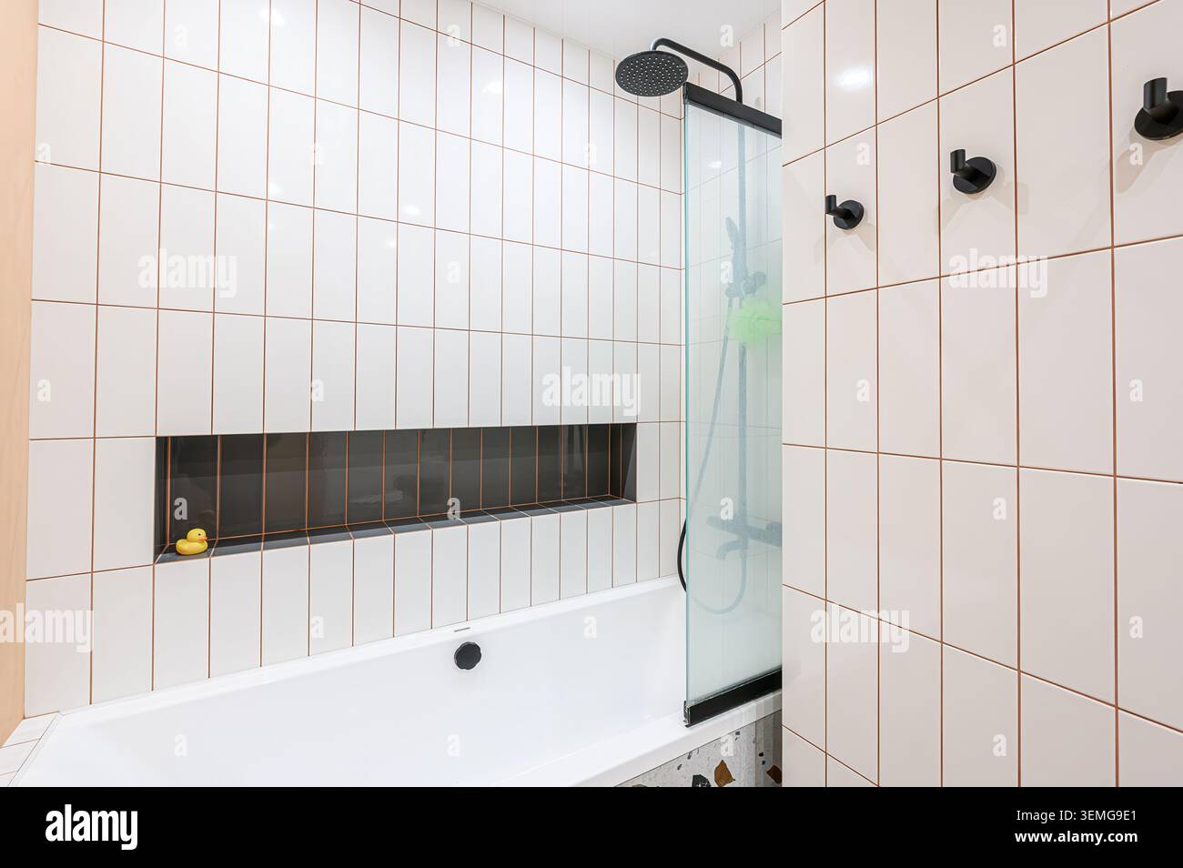 Modernes Bad mit weißen Fliesen, schwarzen Armaturen und einer eingebauten Nische mit einer Gummiente. Badewanne mit Glasdusche. Stockfoto