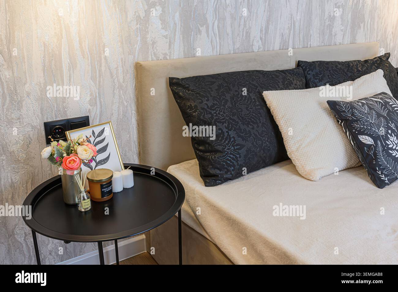 Gemütliche Schlafzimmerecke mit dekorativen Kissen, Nachttisch mit Blumen, Kerzen und gerahmtem Muster. Warmes Licht. Stockfoto
