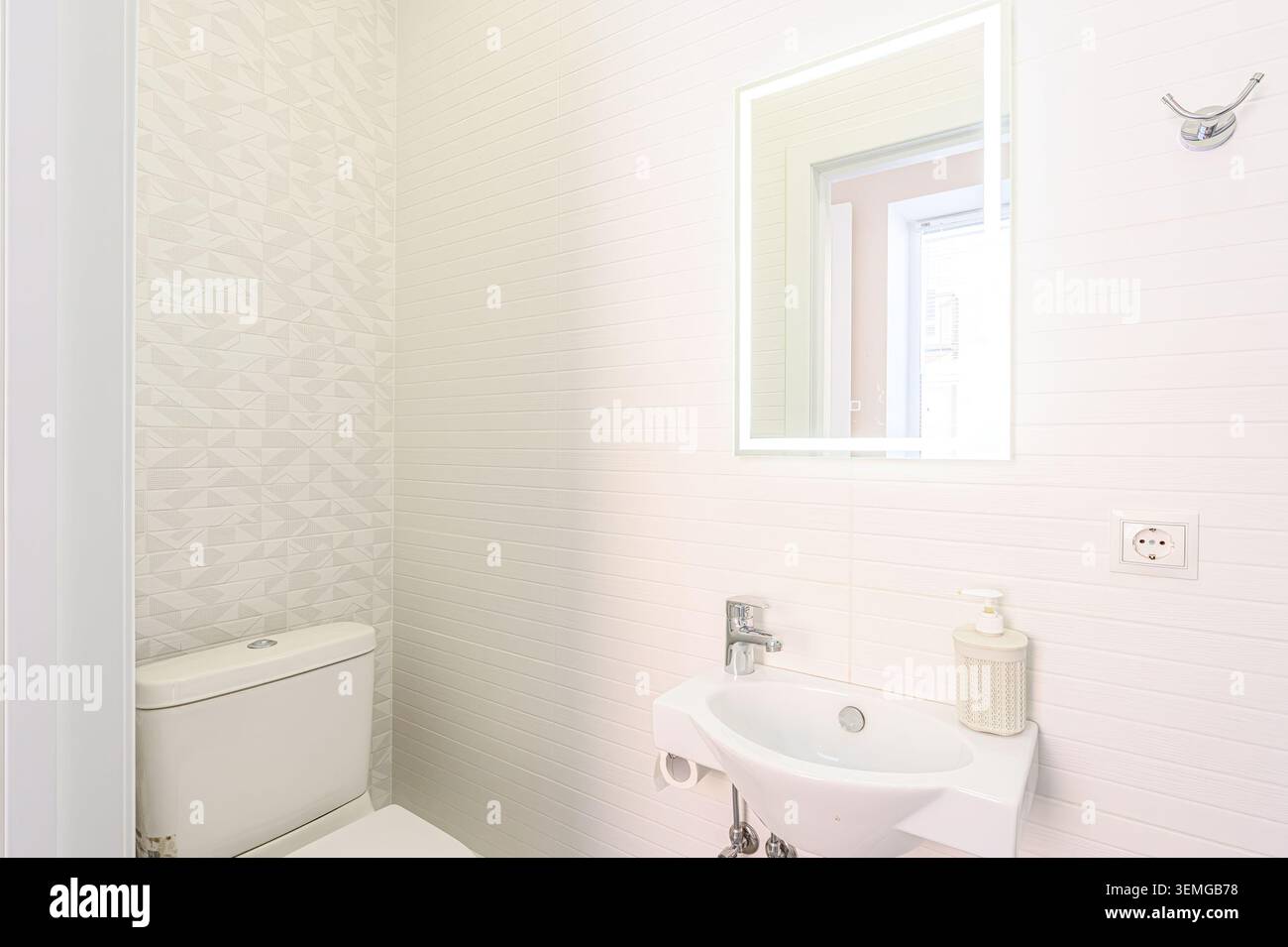 Helles, weißes Badezimmer mit Toilette, Waschbecken und beleuchtetem Spiegel. Die Wände sind mit strukturierten Mustern gefliest. Das gesamte Design ist sauber und modern Stockfoto