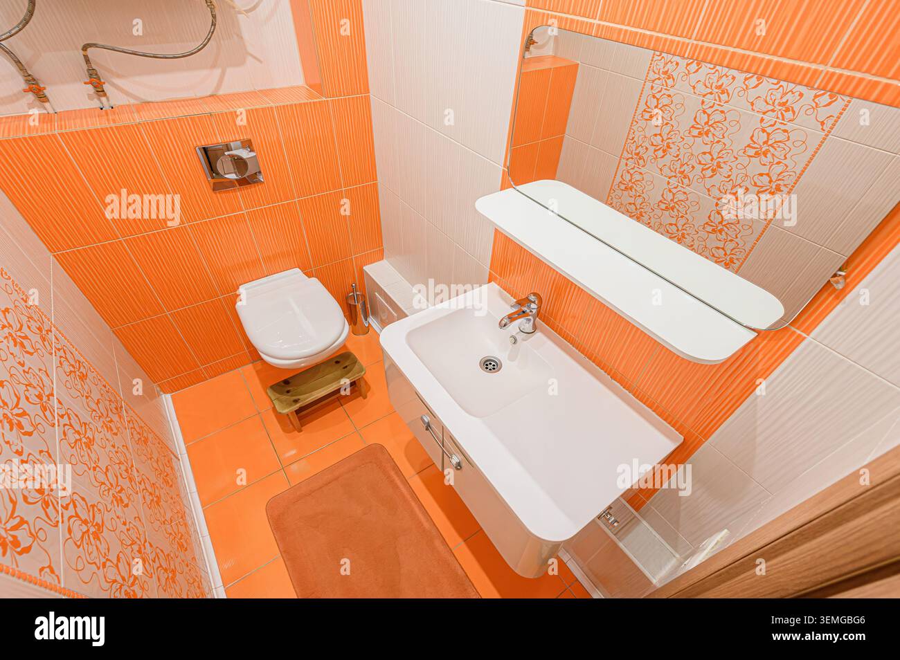 Blick auf die Augenhöhe auf ein kleines Badezimmer mit orangefarbenen und weißen Fliesen, Toilette, Waschbecken, Spiegel und Teppich. Das Farbschema erzeugt ein warmes und lebendiges AT Stockfoto