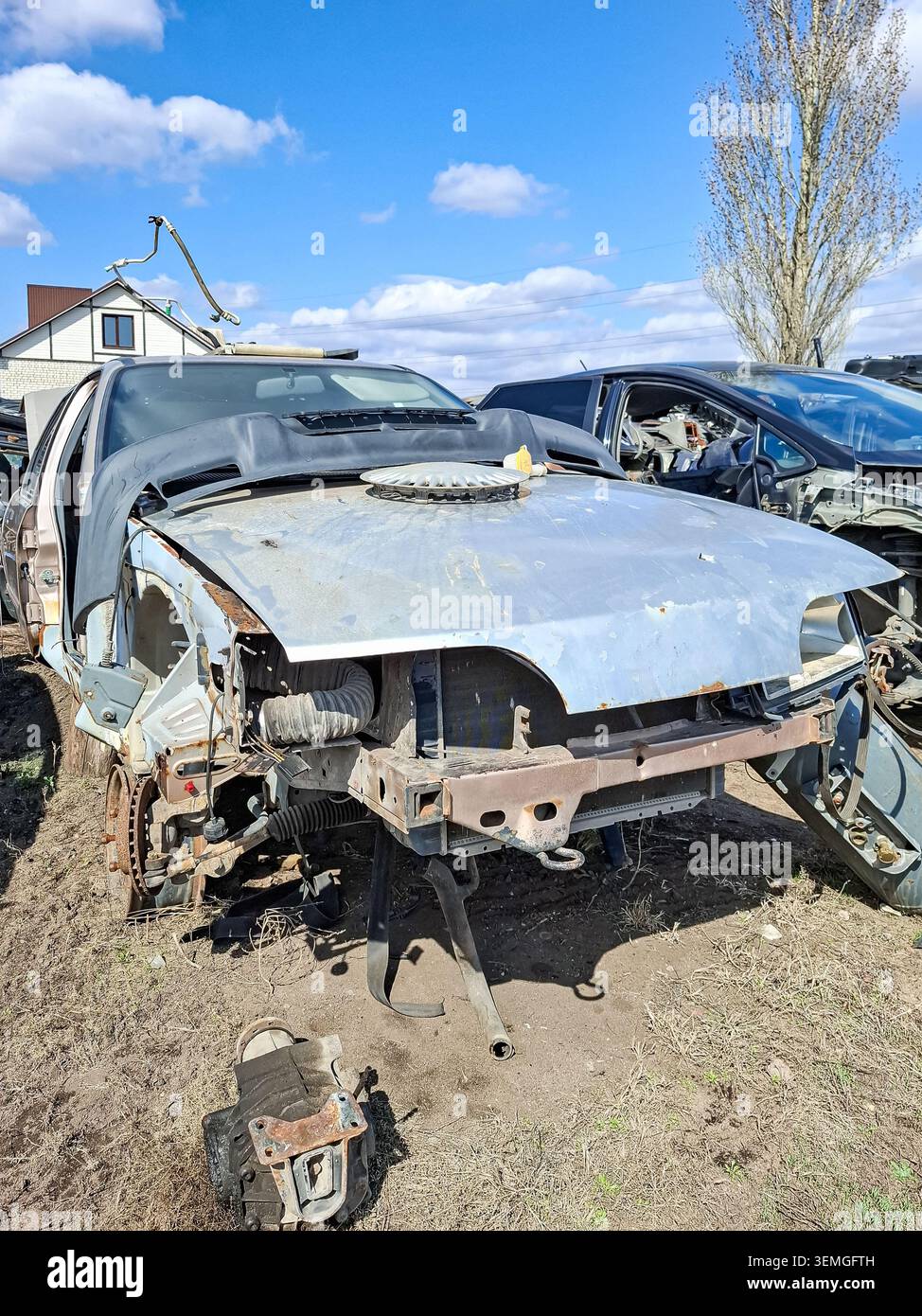 Ein kaputtes silbernes Auto sitzt auf einem Schrottplatz. Der Stoßfänger fehlt und zeigt den Kühler und andere Teile. Fahrzeuge in der Umgebung weisen ebenfalls Anzeichen von Schäden auf. Stockfoto