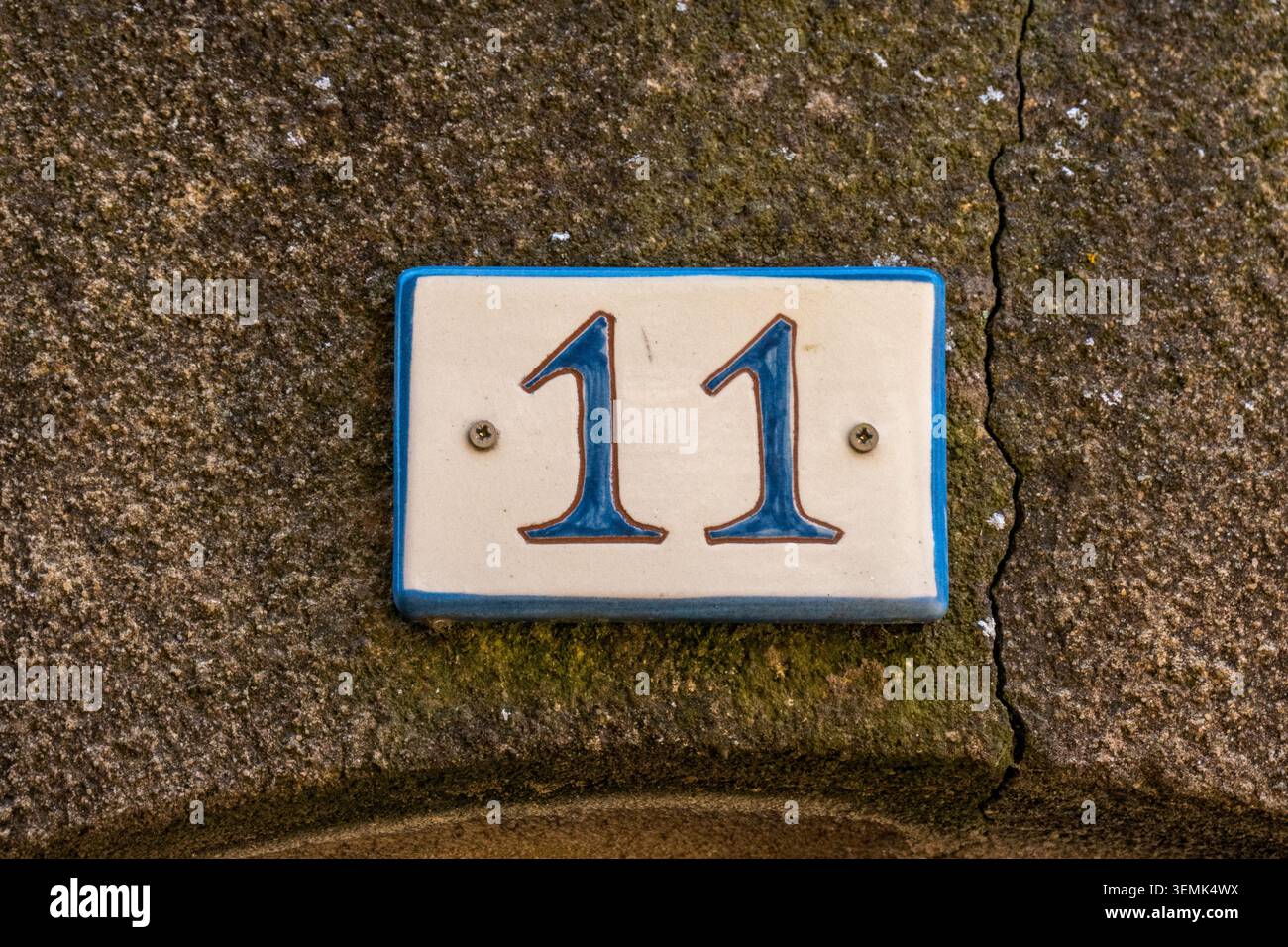 Eine blau-weiße Keramik-Hausnummer 11-Plakette, verschraubt auf eine zerrissene und moosig verwitterte Steinmauer in einem französischen Dorf Stockfoto