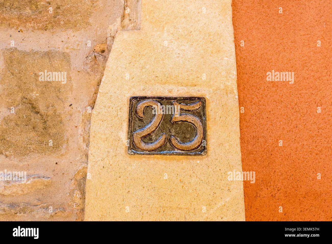 Eine kleine kunstvoll verzierte Bronzetafel mit der Nummer 25, die in einer cremeverzierten Säule neben einer leuchtend orangen Wand in einem französischen Dorf platziert ist Stockfoto