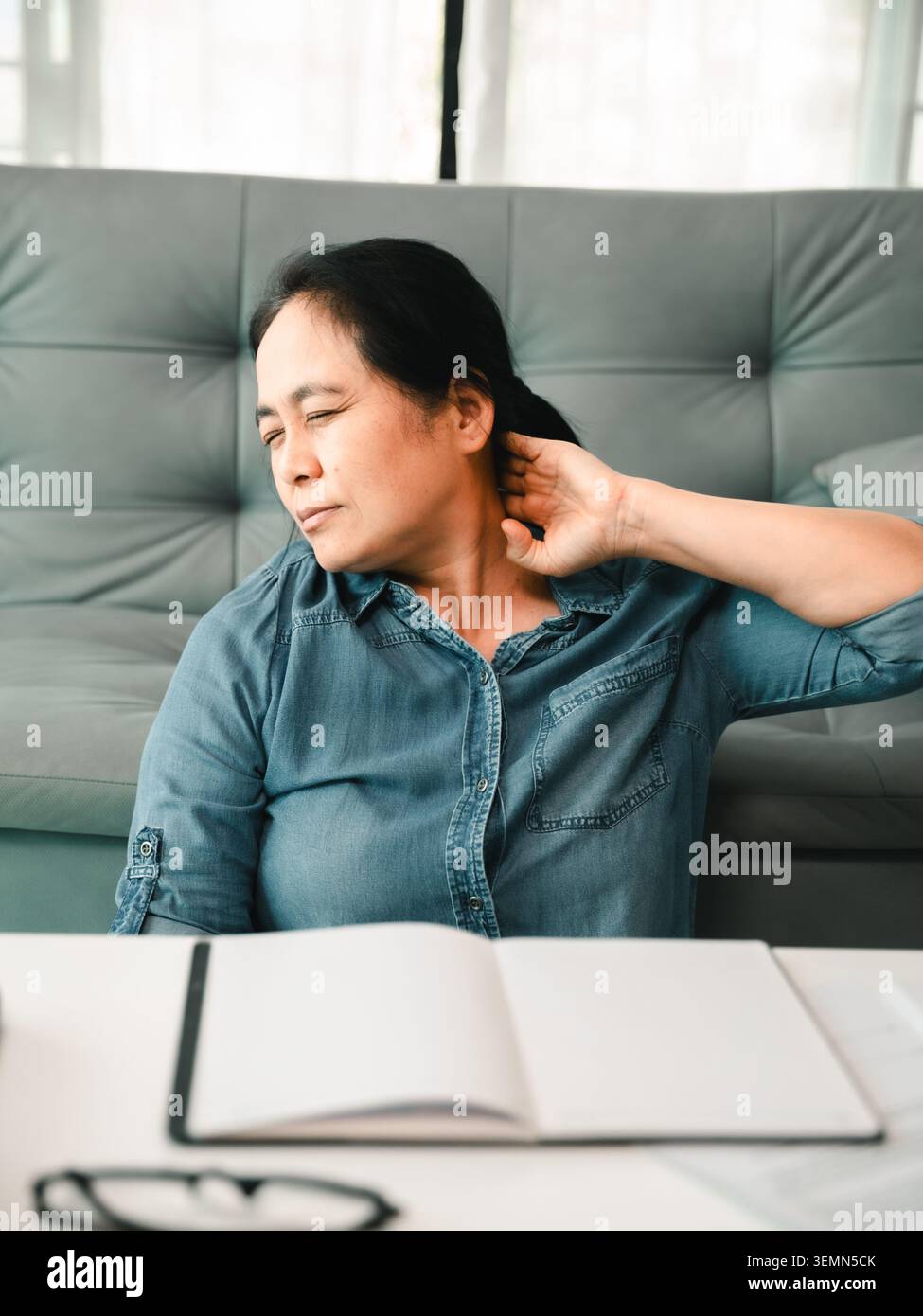 Asiatische Frau mit Nackenschmerzen, die an einem Schreibtisch sitzt, sich neben einem offenen Notizbuch und einer Brille ausdehnt, die Müdigkeit im Büro zeigt, schlechte Körperhaltung, Körperverspannungen, Stockfoto