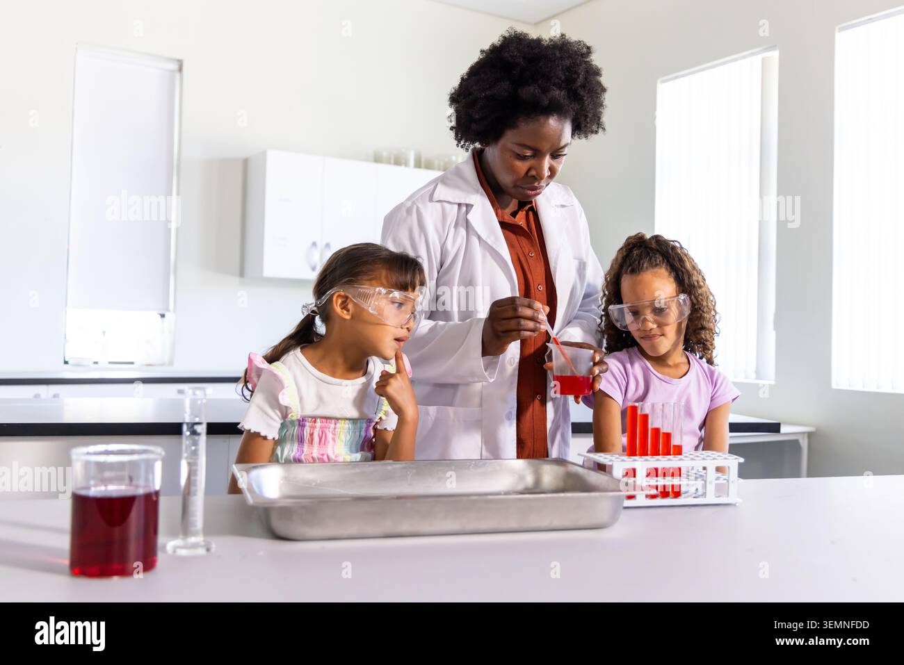 Afroamerikaner-Lehrer im Labormantel, der rote Flüssigkeit in Röhrchen gießt, mit Kindern im Labor Stockfoto