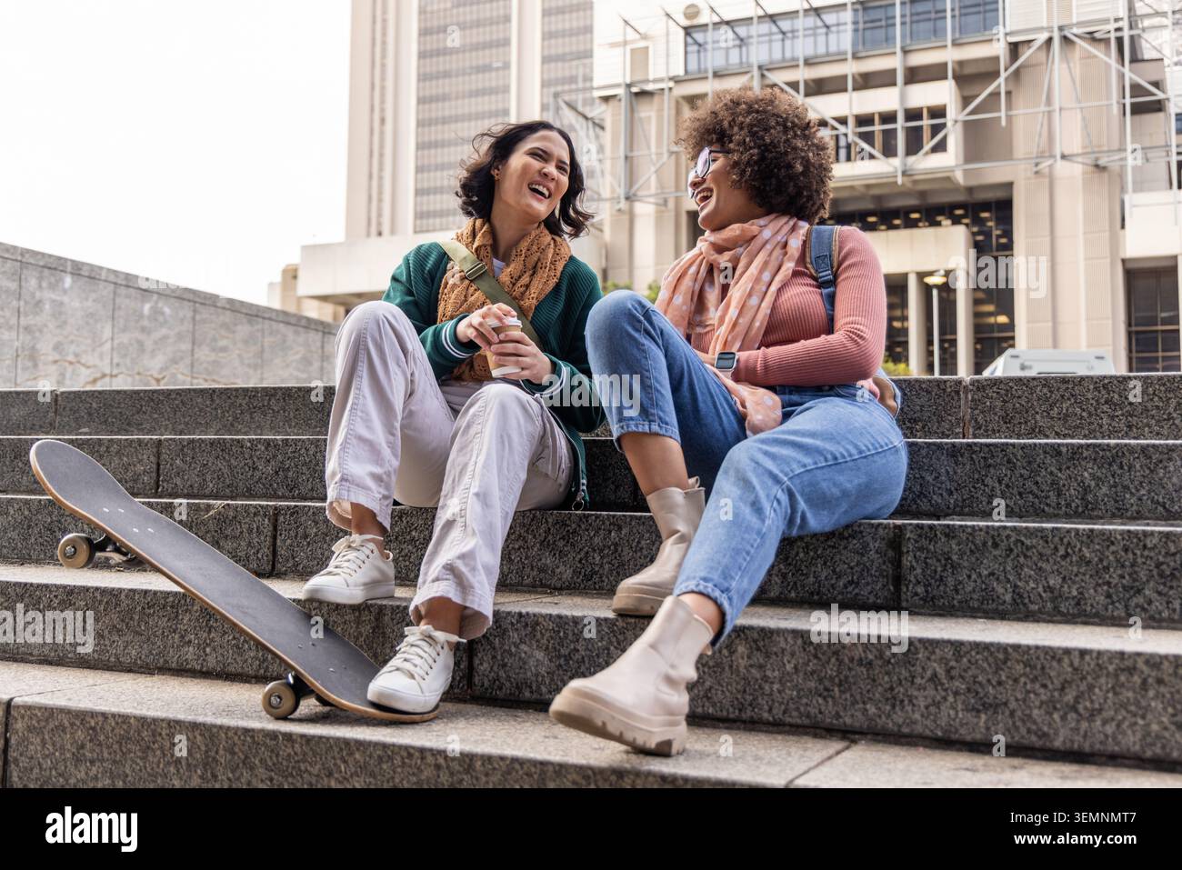 Zwei Freundinnen sitzen auf Steintreppen im städtischen plaza und teilen sich Kaffee, Skateboard und Tücher Stockfoto