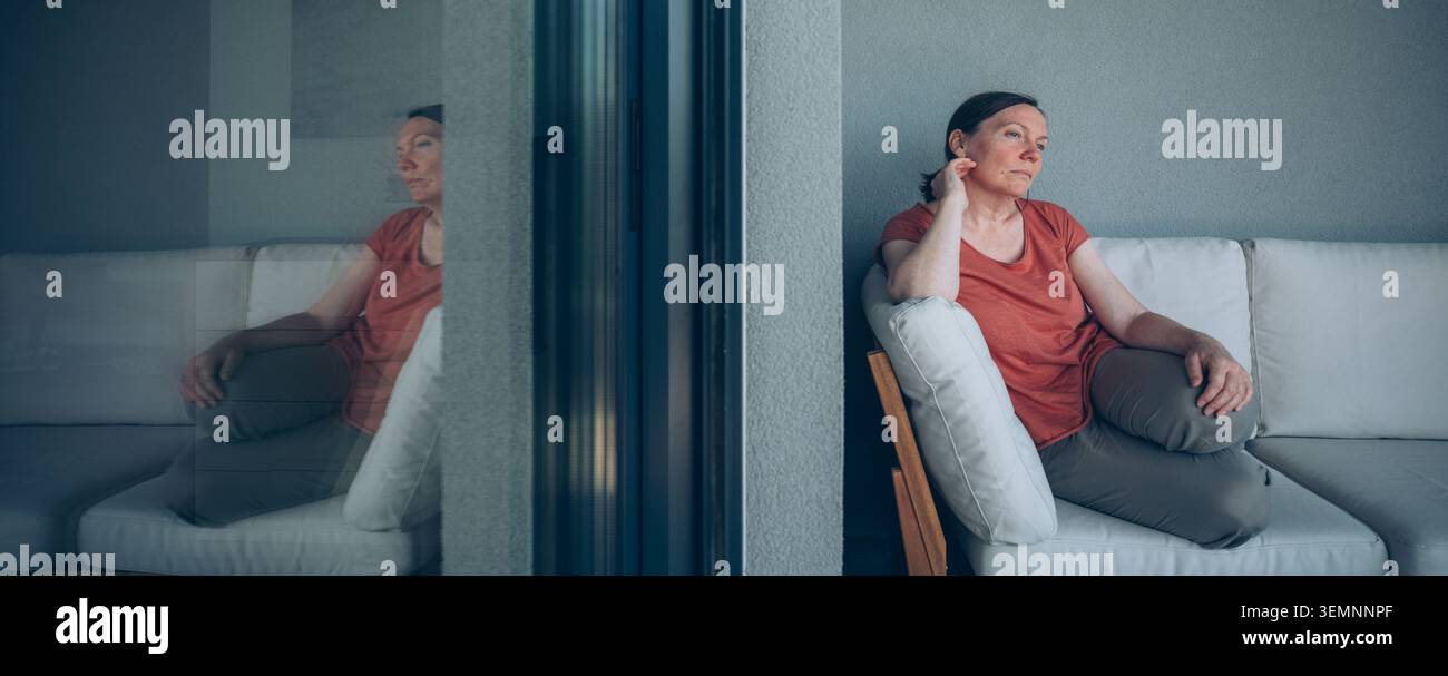 Eine nachdenkliche Frau mittleren Alters sitzt allein auf einer Couch neben einer Glastür und blickt mit einem reflektierenden, melancholischen Ausdruck nach außen Stockfoto