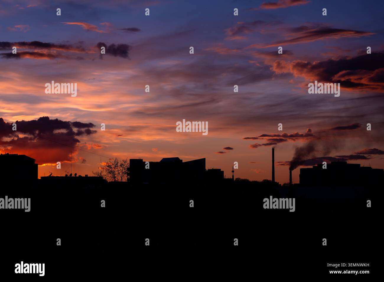 Die Silhouette der Skyline der Stadt bei Sonnenuntergang mit industriellem Rauch, der sich gegen leuchtende orange-violette Wolken erhebt Stockfoto