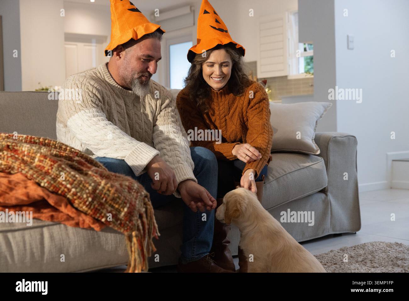 Ein Paar mit Kürbishut sitzt auf dem Sofa und greift zum Welpen, als ein Welpe die Hände drückt und Haustierwerbung macht Stockfoto