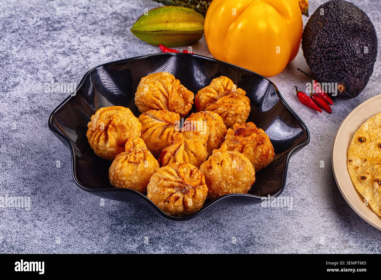 Goldene gebratene momo-Knödel in schwarzer Schüssel mit gelbem Paprika, Avocado, Chili-Paprika, Sternfrucht und Fladenbrot auf Steinoberfläche Stockfoto
