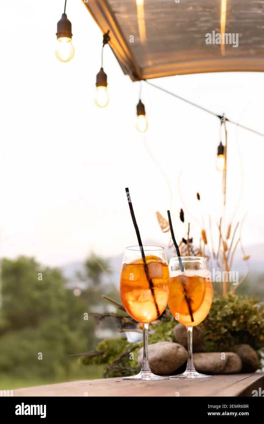 Auf einem rustikalen Holztisch stehen zwei elegante Stielgläser mit leuchtendem Orangencocktail, Eiswürfeln und schwarzen Strohhalmen Stockfoto
