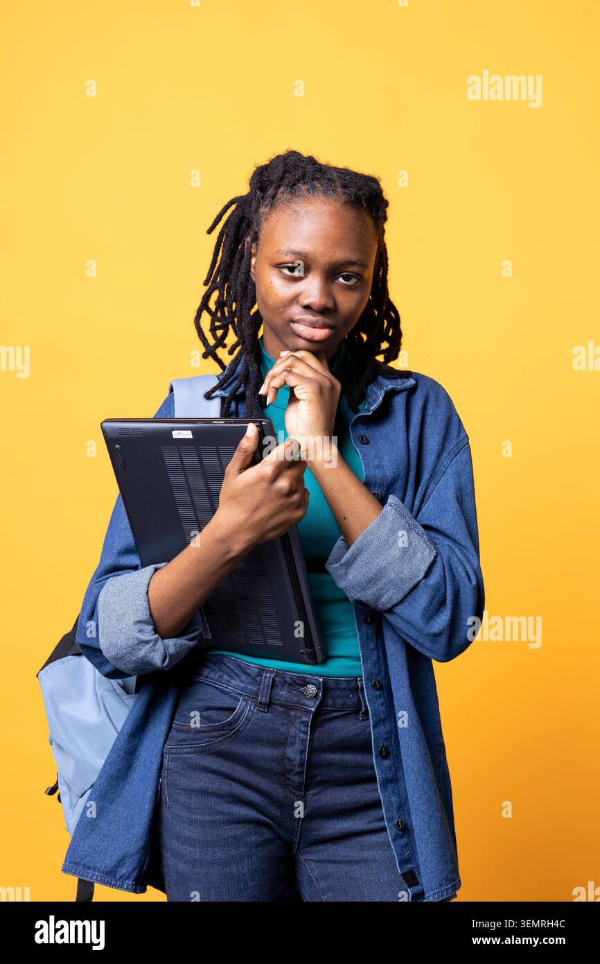 Porträt einer BIPOC nachdenklichen Frau mit Laptop, die es benutzt, um Informationen für die Prüfung, Studiohintergrund, afroamerikanischer Student mit Notebook, der sich vor der Universitätsbewertung an Informationen erinnert Stockfoto