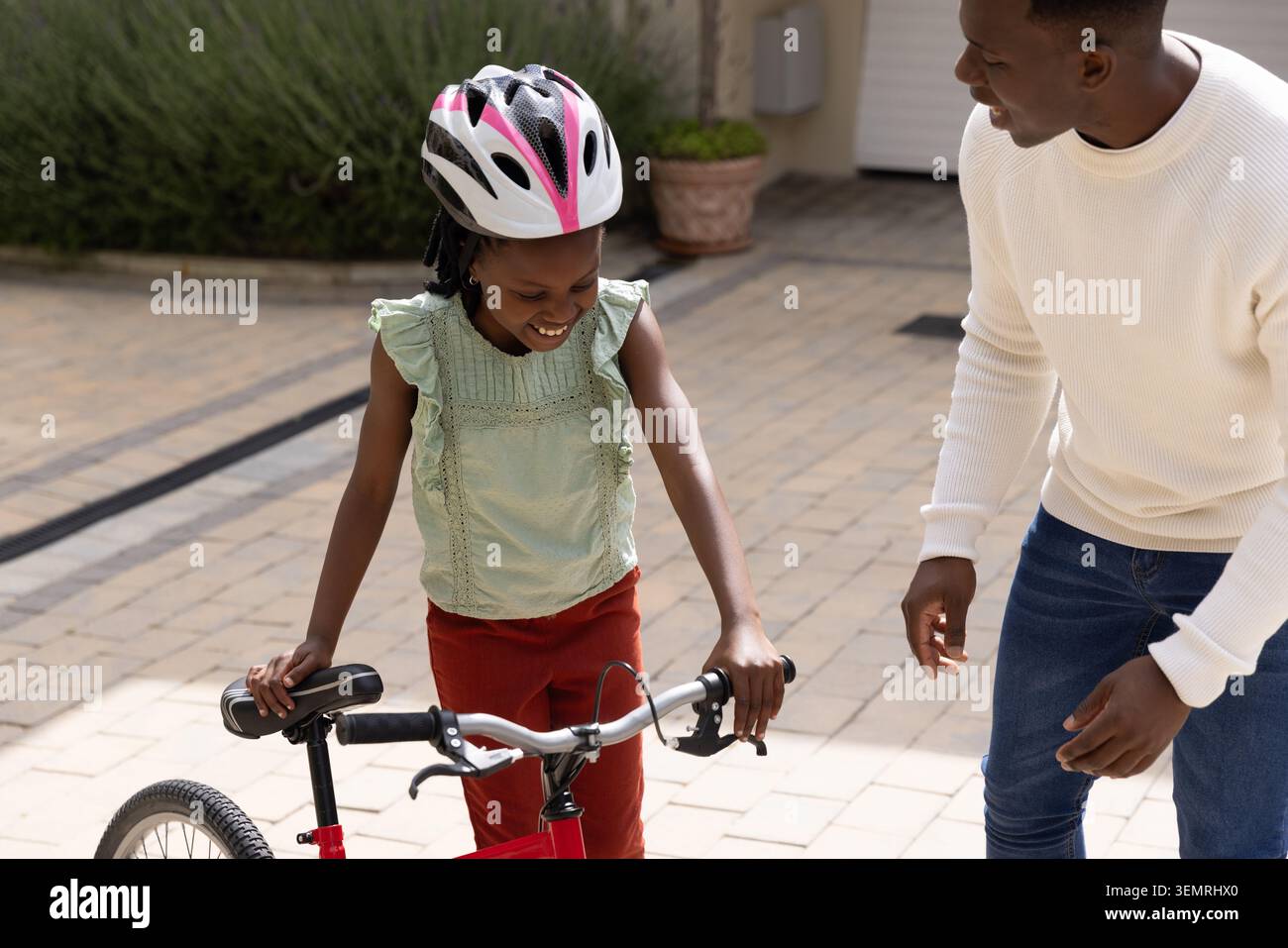 Afroamerikanischer Vater und Tochter üben auf rotem Fahrrad auf einer gepflasterten Einfahrt mit Helm Stockfoto