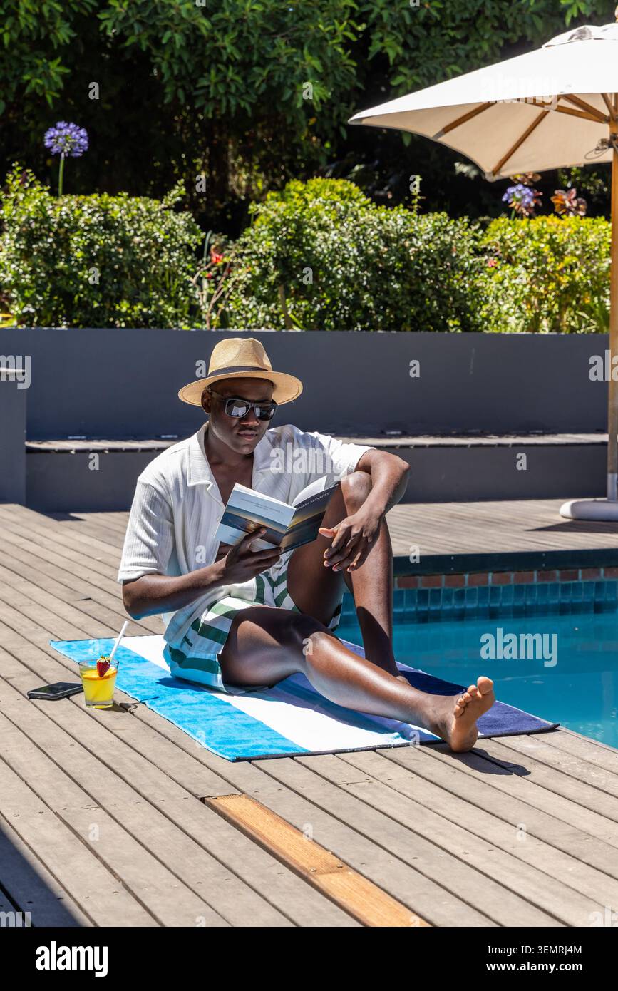 Afroamerikaner, der auf dem Pooldeck sitzt, Handtuch liest Taschenbuch mit Strohhut und Sonnenbrille Stockfoto