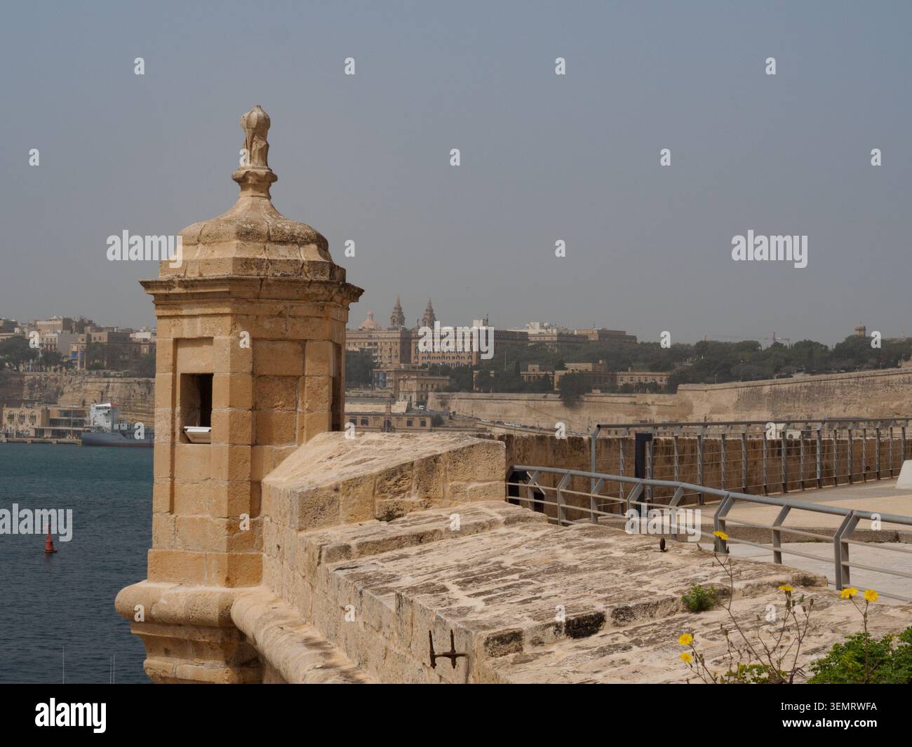 Die Uferpromenade von Birgu mit Valletta, die an einem sonnigen Tag über das Wasser sichtbar ist. Fort St. Angelo im Vordergrund. Stockfoto